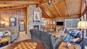 A cozy cabin interior with a stone fireplace, wooden walls, and a large window overlooking a deck. - 4BR Lakehouse | Sleeps 10 | Fire Pit, Dock, Kayaks (Long Pond)