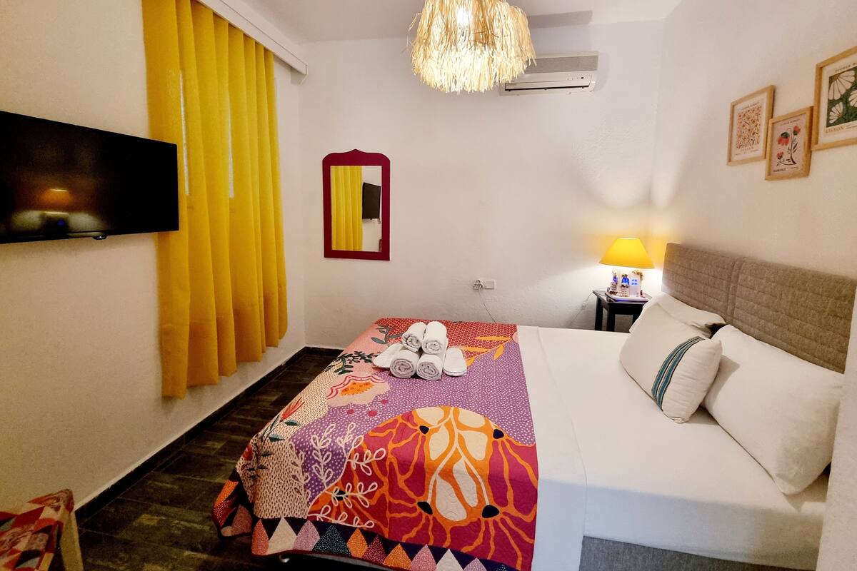 Una habitación de hotel con cama, televisión, espejo, lámpara y ventilador de techo.