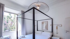 A modern bedroom with a four-poster bed, a bedside table, a wall-mounted lamp, and a window with curtains. - Colosseum Cosy Apartment Close To Metro (Rome)