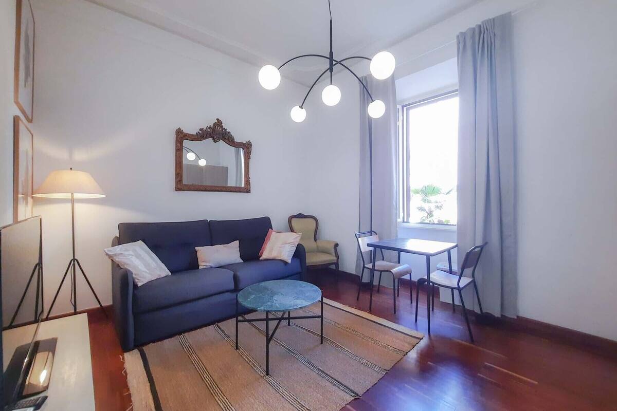 Un salon avec un canapé bleu foncé, une table basse ronde, une petite table avec des chaises, un lampadaire, un miroir et une grande fenêtre avec des rideaux.