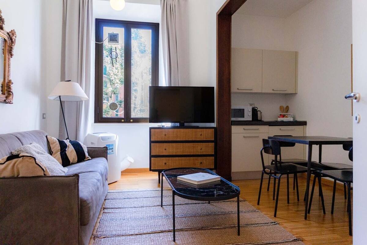 Un espace de vie compact comprenant un canapé, une table basse, un téléviseur, un coin cuisine avec des placards et un four à micro-ondes, ainsi qu’une fenêtre avec des rideaux.