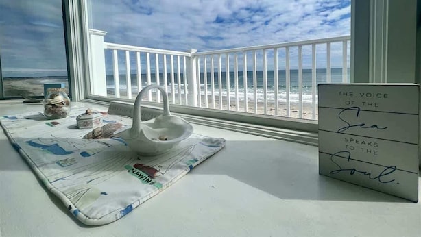 A room with a view of the beach, a white table with a mat, a decorative bowl, a jar, and a sign with a quote. - Oceanfront Wells Beach Condo - Ocean Surf #5 (Wells)