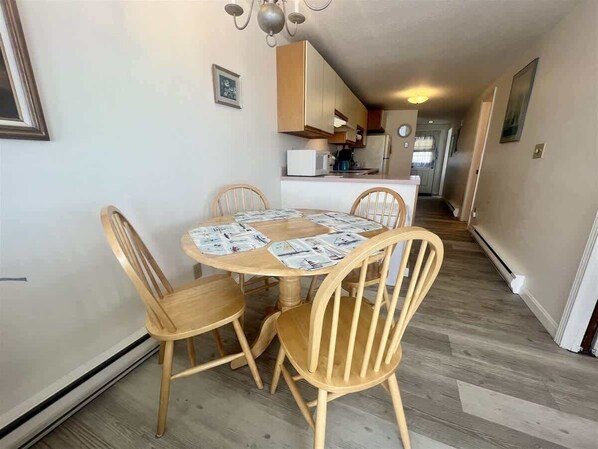 A dining area with a round table and four wooden chairs, a kitchen in the background, and a hallway leading to another room.