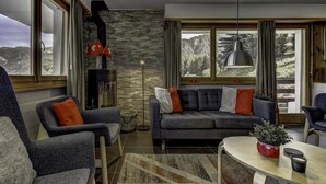 A modern living room with a stone wall, a fireplace, a sofa with orange cushions, and a view of a mountainous landscape through large windows. - Appartement Mont Parrain 4 - La Tzoumaz, 4 Vallees (La Tzoumaz)