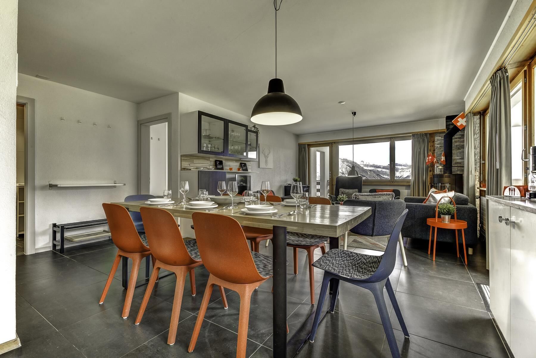 A modern kitchen with a dining area, featuring a large island, orange and blue chairs, and a view of a snowy landscape through the windows.