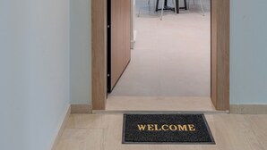 A hallway with a wooden door leading to a dining area with a white table and chairs, a red refrigerator, and a welcome mat on the floor. - Harbour Hill Suite 4—200 m. away from the beach (Rethymno)
