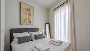 A neatly made double bed with pillows, a bedside table with a lamp, and a large window with curtains. - Harbour Hill Suite 4—200 m. away from the beach (Rethymno)