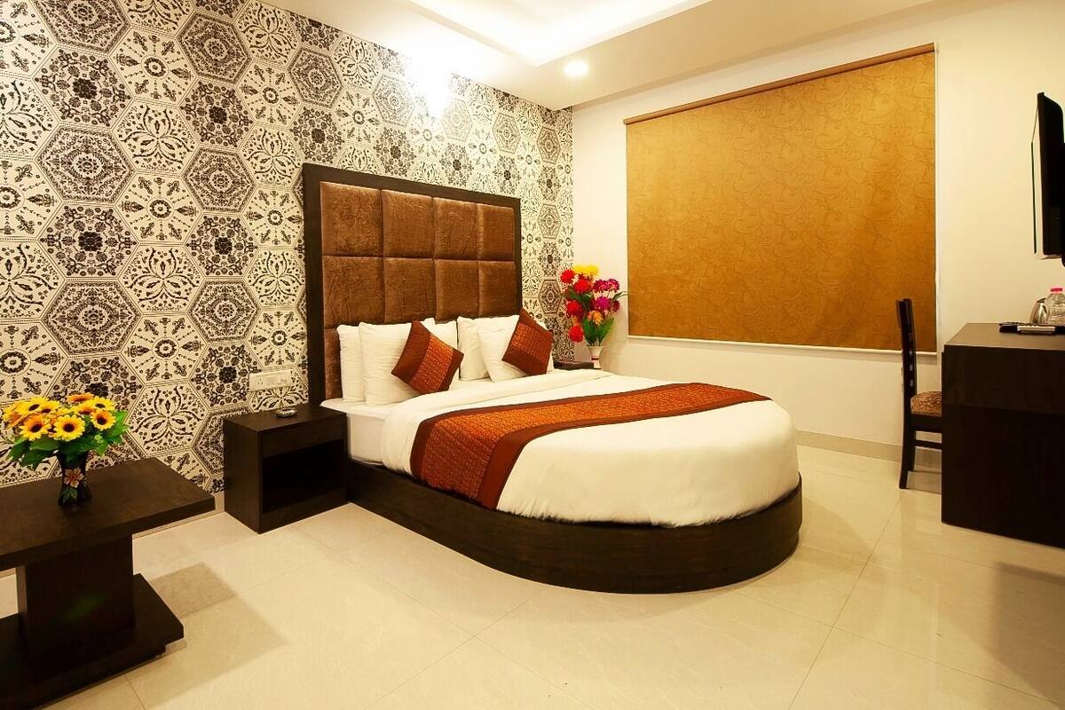 Una habitación de hotel moderna con una cama curvada, papel tapiz estampado, un ventanal con persiana, una pequeña mesa con un jarrón de flores y un televisor sobre un soporte.
