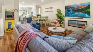 En moderne stue med en stor sofa, et lille rundt bord og en pejs. I baggrunden er der et spiseområde med et køkken.