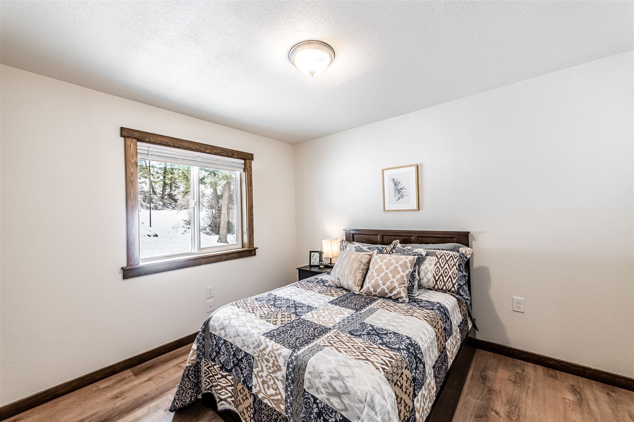 A bedroom with a wooden floor, a bed with patterned bedding, a nightstand with a lamp, a window with a view of trees, and a ceiling light.
