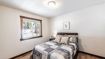 A bedroom with a wooden floor, a bed with patterned bedding, a nightstand with a lamp, a window with a view of trees, and a ceiling light.