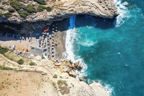 Vue aérienne d’une zone côtière avec une plage, des falaises rocheuses et un petit bâtiment doté d’un toit rouge.