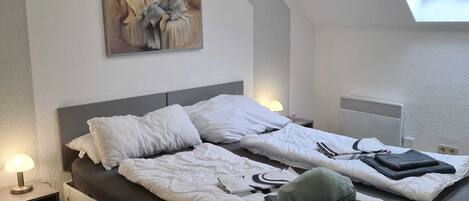 Un dormitorio con cama, un cuadro, tragaluz, mesita de noche y calefactor.