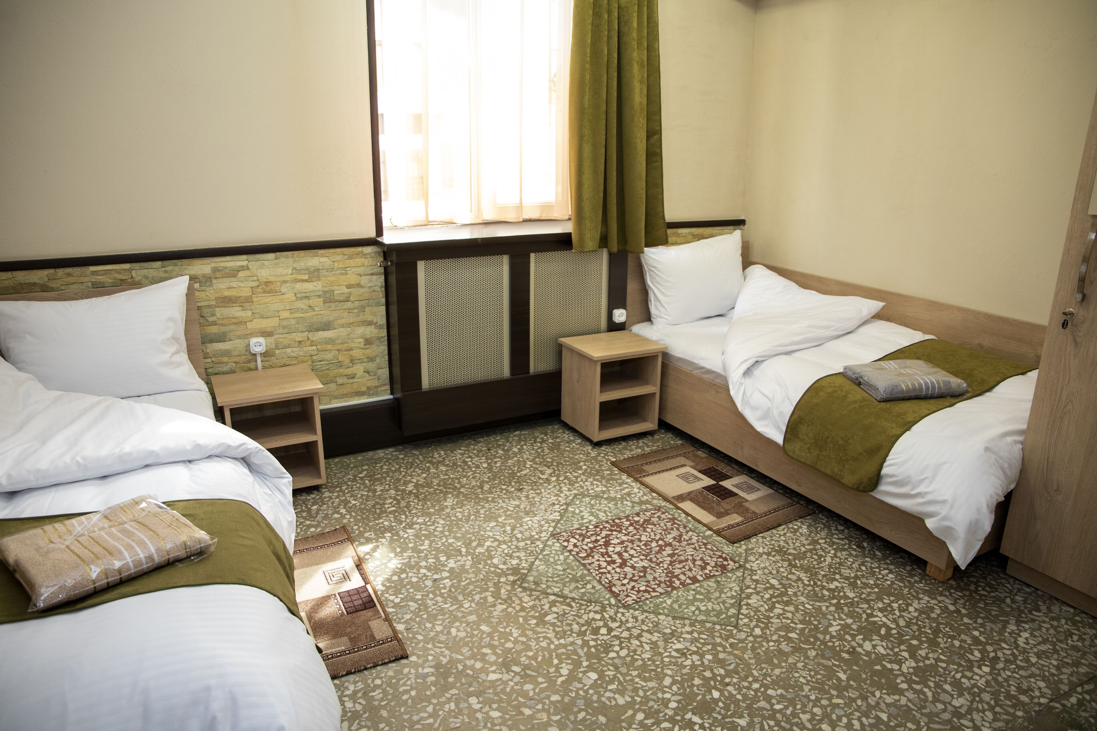 A hotel room with two beds, a window with curtains, a radiator, and a small wooden nightstand.