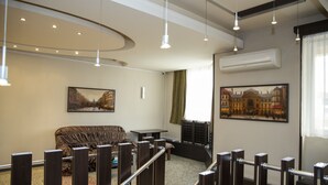 Un hall d’hôtel avec un canapé marron, une petite table et un tableau représentant un bâtiment. On y trouve des luminaires au plafond et un appareil de climatisation.