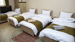 Une chambre d’hôtel avec quatre lits alignés, chacun doté de draps blancs et d’une tête de lit marron. Une petite table se trouve entre les lits, et un plaid vert est posé sur le sol.