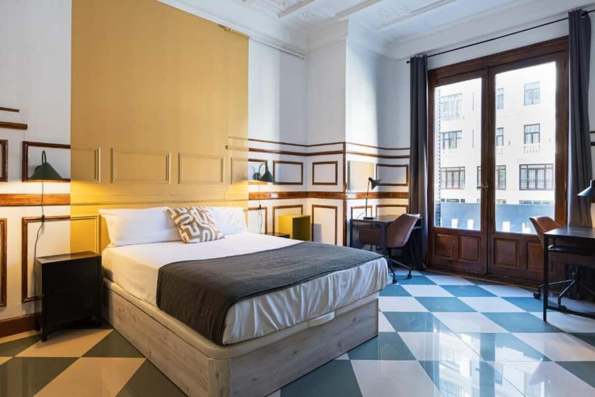 A hotel room with a large bed, a desk, a chair, and a view of the city through large windows.