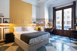 A hotel room with a large bed, a desk, a chair, and a view of the city through large windows. - Live It Gran Via (Madrid)