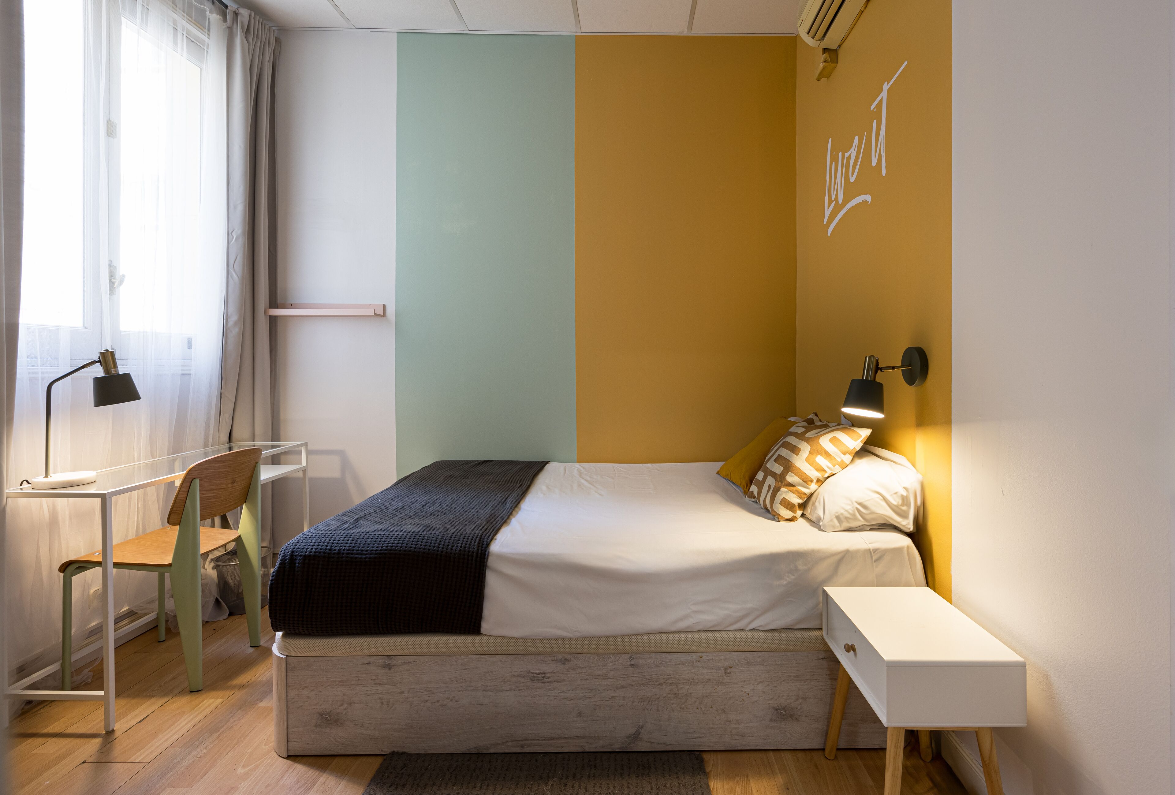 Een slaapkamer met een bed, nachtkastje, lamp, stoel en bureau.