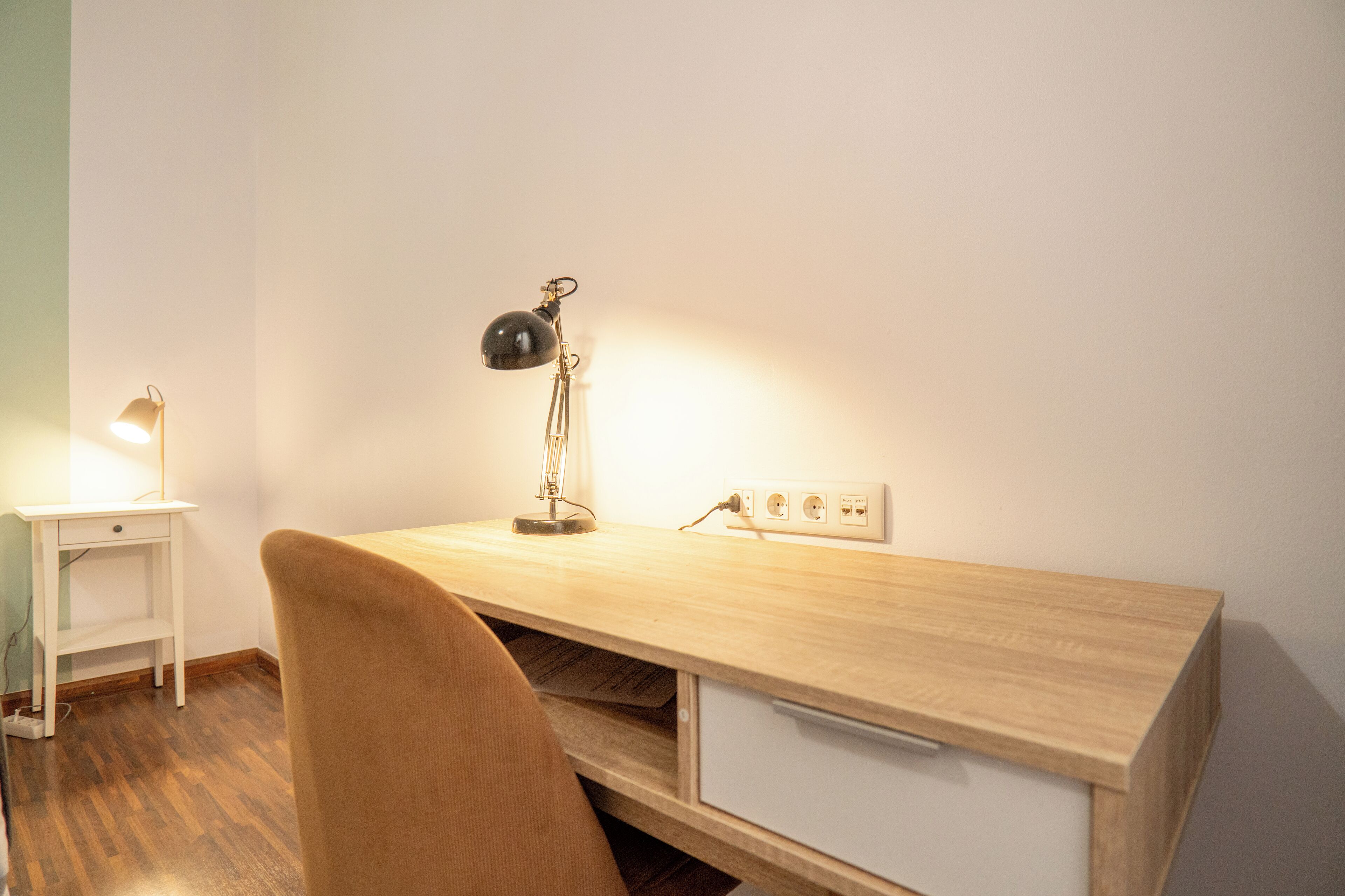 A small, neatly organized workspace with a wooden desk, a chair, a lamp, and a power outlet.