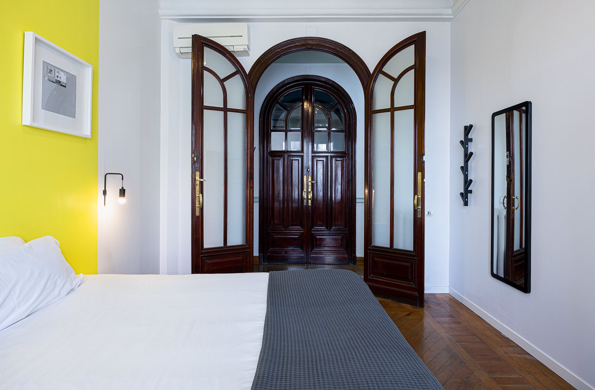 Quarto de hotel com cama, porta de madeira com painéis de vidro, luminária fixada na parede e quadro emoldurado.
