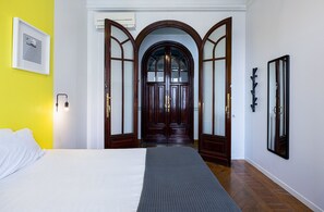 A hotel room with a bed, a wooden door with glass panels, a wall-mounted light fixture, and a framed picture.