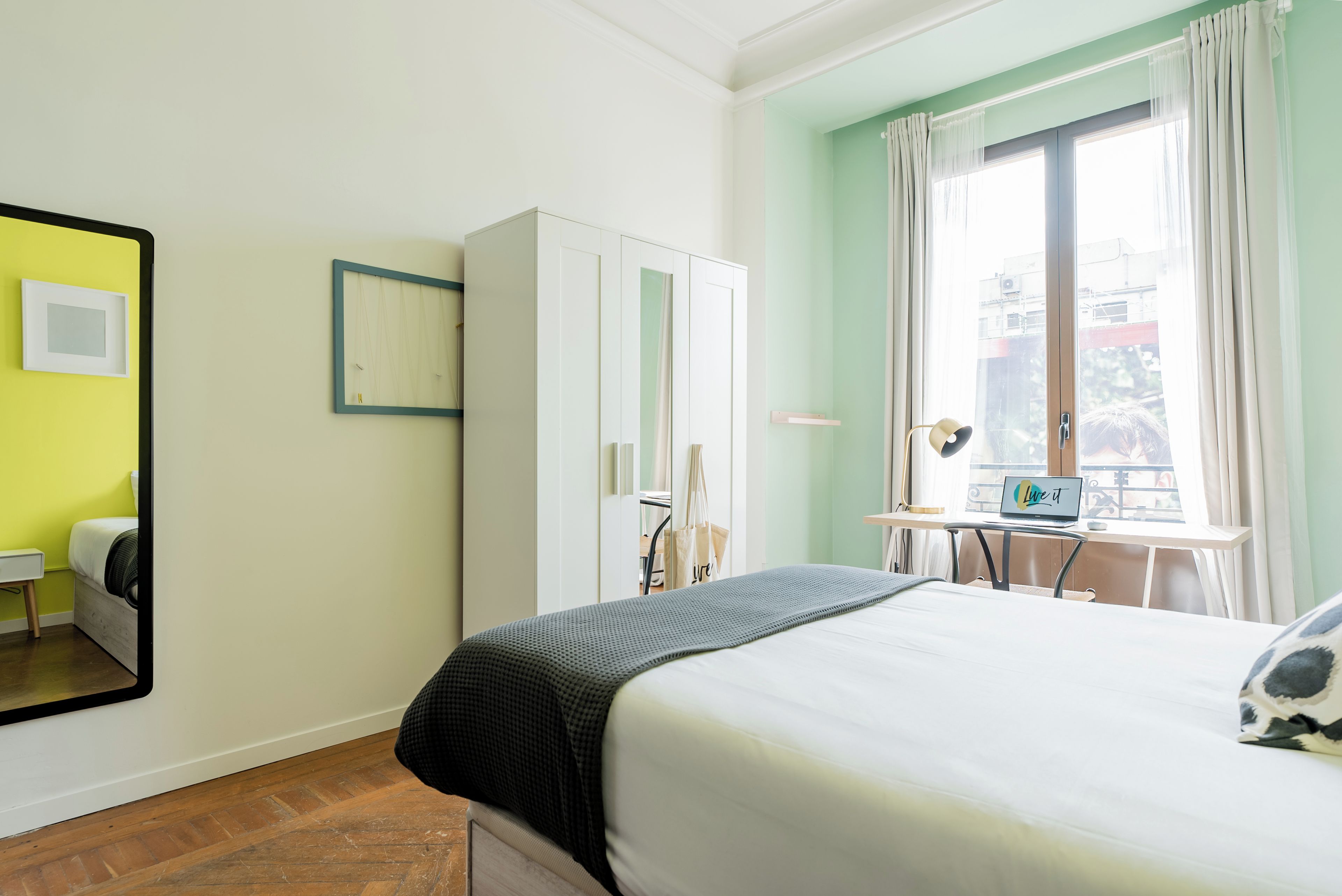 Ein Schlafzimmer mit Bett, Schreibtisch, Laptop, Kleiderschrank und einem Fenster mit Vorhängen.