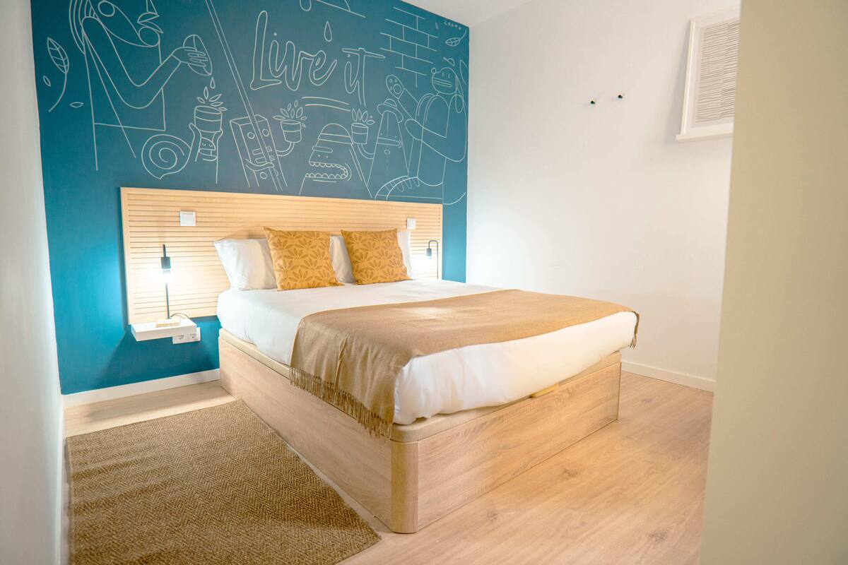 A bedroom with a wooden headboard, a bed with white and beige bedding, and a wall mural.