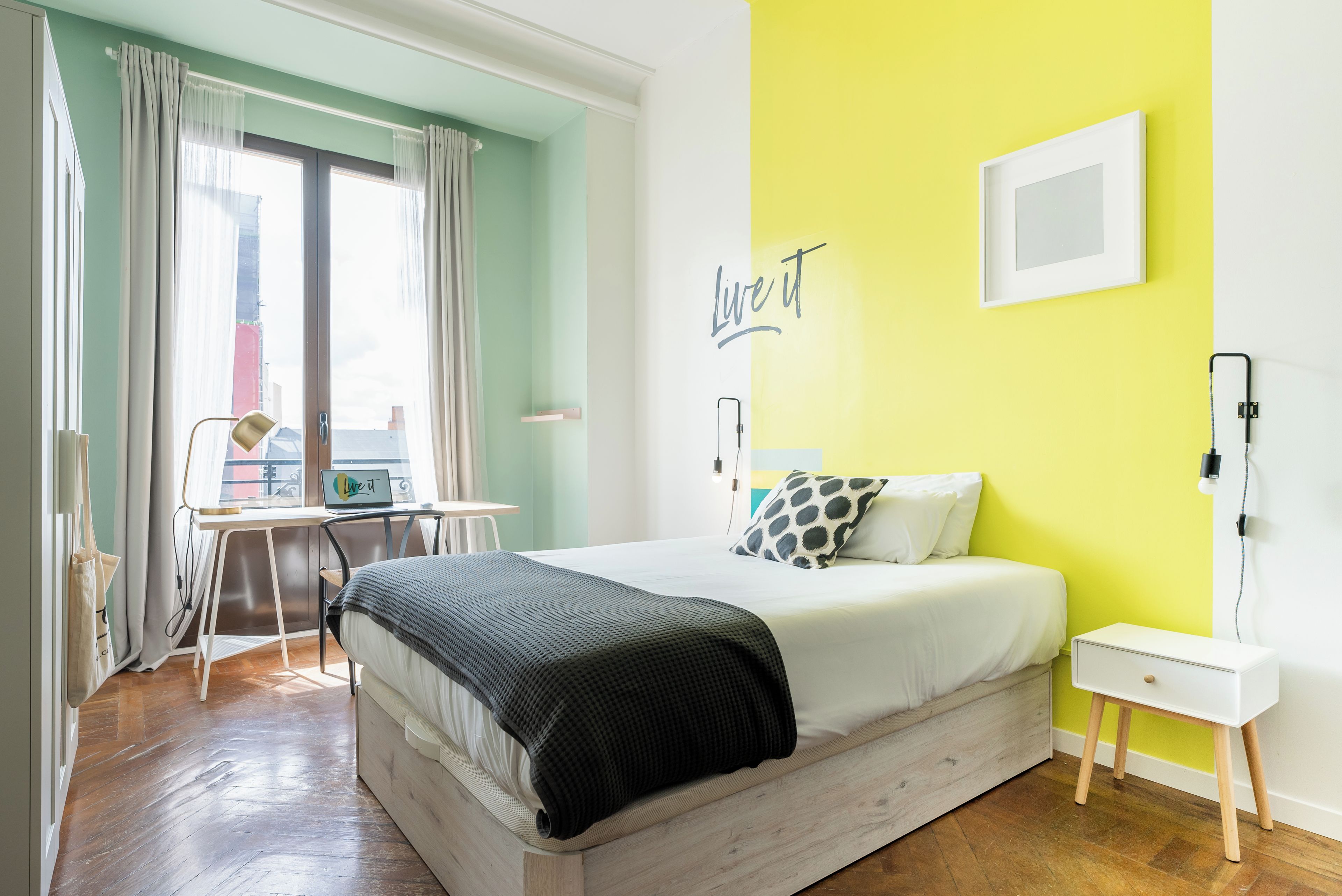 A bedroom with a bed, a desk, and a chair. There is a window with curtains, a wall with a 'Live it' sign, and a bedside table with a lamp.