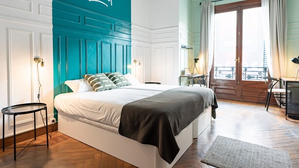 A bedroom with a large bed, a teal accent wall, a wooden floor, and a balcony with a view. - Live It Gran Via (Madrid)