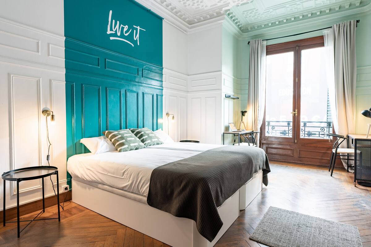 A bedroom with a large bed, a teal accent wall, a wooden floor, and a balcony with a view.