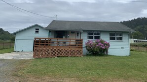 A light blue single-story house with a porch, a wooden fence, and a small garden with pink flowers.