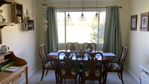 A dining area with a wooden table and chairs, a window with curtains, and a shelf with decorative items.