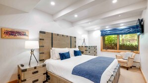 A bedroom with a large bed, a bedside table with a lamp, a chair, and a window with a view of greenery.