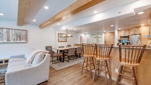 A modern kitchen with wooden cabinets, a bar area with rustic stools, and a dining area with a grey sofa and a wooden coffee table.