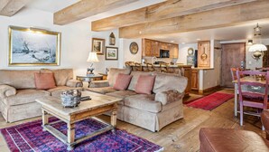 Ein großzügiges Wohnzimmer mit einer großen Couch, einem Couchtisch und einem Essbereich mit Tisch und Stühlen. Im Raum sind Holzbalken unter der Decke und im Hintergrund eine Küche zu sehen.