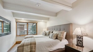 A bedroom with two beds, a nightstand, a lamp, and a window with a view of trees. - Lodge at Lionshead 203 (Vail)