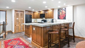 A kitchen with wooden cabinets, a granite countertop, a refrigerator, a microwave, and a painting on the wall. - Lodge at Lionshead 203 (Vail)