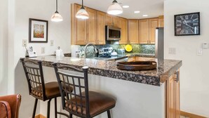 A kitchen with granite countertops, wooden cabinets, a stainless steel refrigerator, a microwave, and a clock on the wall. - Lodge at Lionshead 204 (Vail)