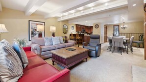 A spacious living room with a red sofa, a grey sofa, and a wooden coffee table. There is a dining area with a table and chairs, and a kitchen in the background. - Lodge at Lionshead 208 (Vail)
