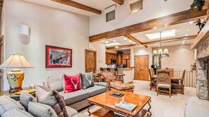 A spacious living room with a sofa, armchairs, and a coffee table. There is a fireplace, a chandelier, and a kitchen in the background. - Lodge at Lionshead 404 (Vail)