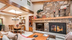 A spacious living room with a stone fireplace, wooden beams, a chandelier, and a dining area with a wooden table and chairs. - Lodge at Lionshead 404 (Vail)