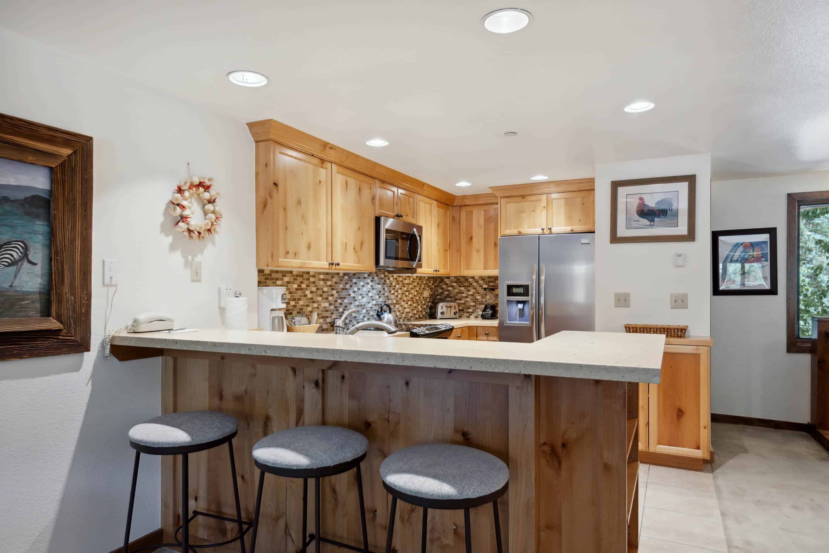 A modern kitchen with wooden cabinets, a stainless steel refrigerator, a microwave, and a bar area with stools.