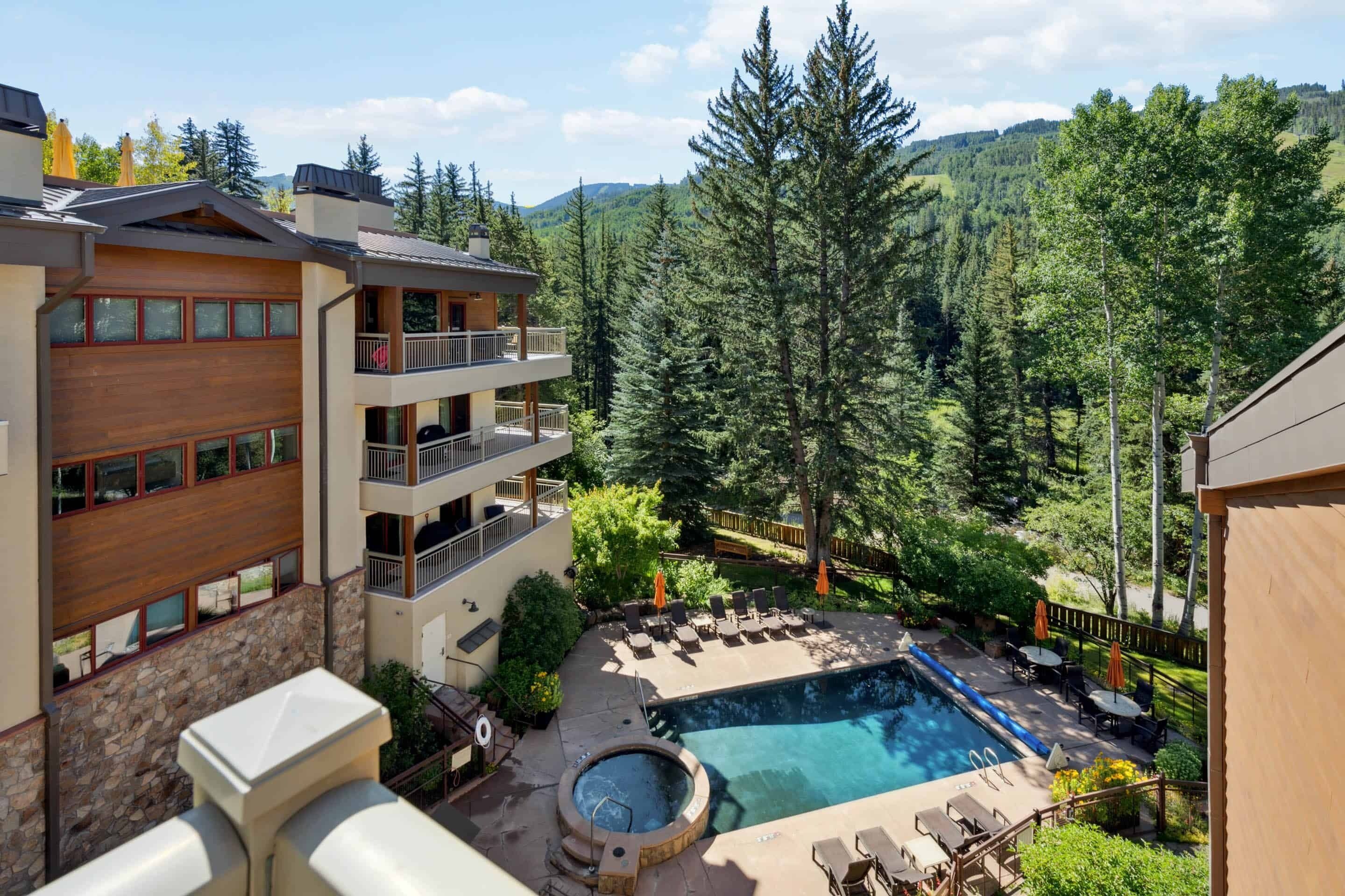 Un edificio moderno con piscina, mobiliario exterior y vistas a una montaña boscosa.