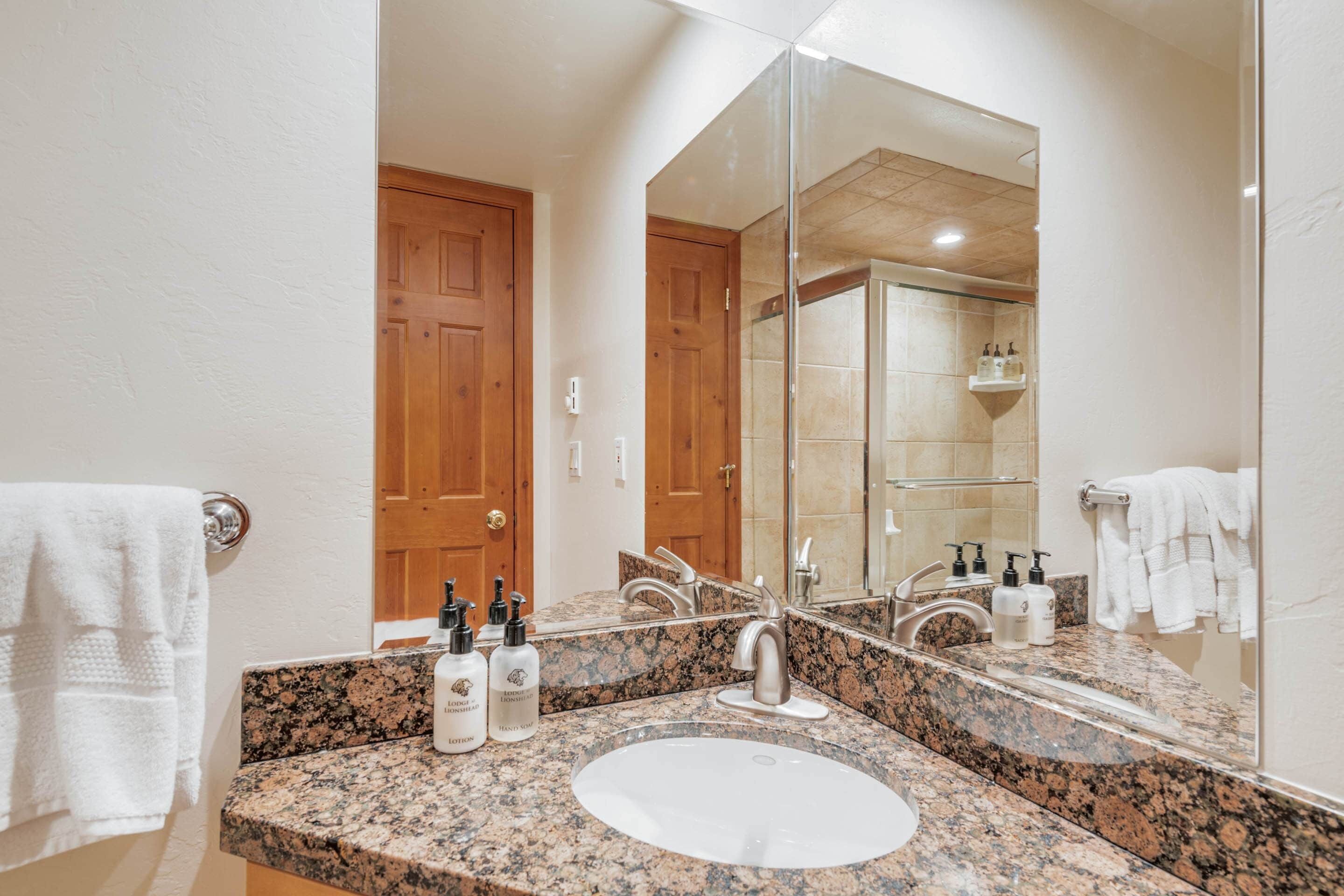 A bathroom with a granite countertop, a white sink, and a large mirror. There are two wooden doors and a shower visible in the reflection.