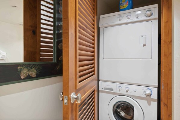 A laundry area with stacked washers and dryers, a wooden door with a glass panel, and a pine cone decoration.