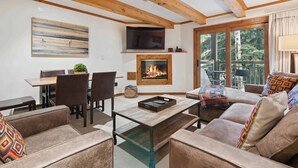A cozy living room with a fireplace, comfortable seating, a dining area, and a large window offering a view of the outdoors. - Lodge at Lionshead 120B (Vail)