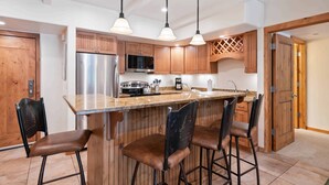 A modern kitchen with wooden cabinets, a stainless steel refrigerator, a microwave, and a coffee maker. There are three bar stools and pendant lights above the island. - Lodge at Lionshead 150A (Vail)