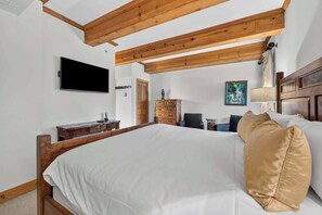 A bedroom with a large bed, wooden ceiling beams, a flat-screen TV, a dresser, and a painting on the wall.