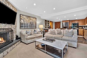 A living room with a fireplace, sofas, a coffee table, and a kitchen area with wooden cabinets and appliances. - Lodge at Lionshead 210 (Vail)